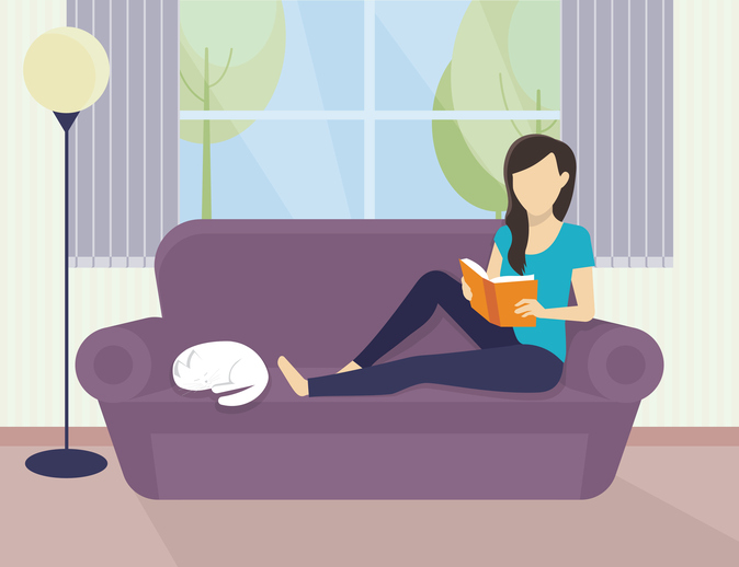 Female sitting on a couch reading a book next to a cat